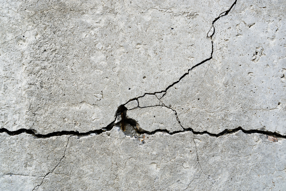 Fissure linéaire traversant un dallage béton industriel gris, vue rasante mettant en évidence le défaut, environnement d'entrepôt logistique