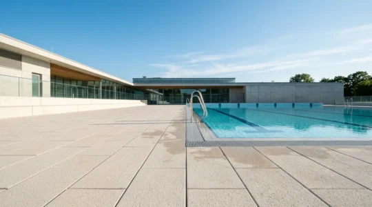 Plage de piscine publique municipale avec dallage antidérapant clair, nombreux baigneurs, environnement estival